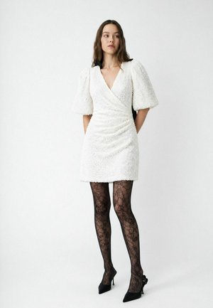 Femme portant une robe portefeuille blanche texturée avec des manches ballon, des collants noirs à motif dentelle et des chaussures à talons noirs, debout devant un fond uni.