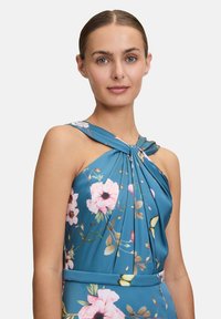 Blumenkleid aus türkisfarbenem Stoff mit gefälteltem Design, verziert mit pinkfarbenen und weißen Blumen, hat einen gedrehten Ausschnitt und eine taillierte Passform.