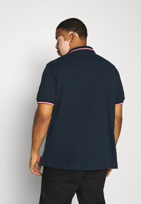 Navy polo-skjorte med korte ermer, med en kontrasterende rød og hvit stripe på kragen og ermeender. Glatt bomullsstruktur.