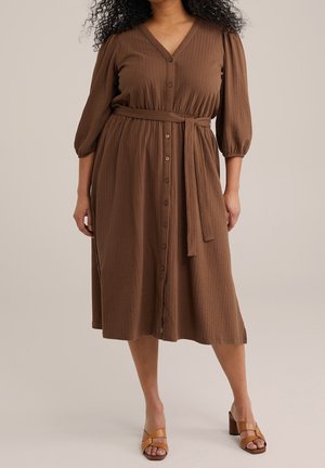 Kvinde iført en brun midi-kjole med knapper, taljebånd og trekvart pufærmer, kombineret med brune åbne block-hel sandaletter.