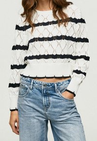 Woman wearing a white knitted cropped sweater with black horizontal stripes and light blue high-waisted jeans, one hand in pocket.