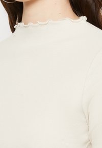 Långärmad topp i ribbat tyg, krämfärg, med en hög hals och en volangdetalj. Närbild av axeldesign.