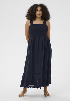 Vrouw met krullend haar die een navyblauwe mouwloze midi-jurk draagt met een gesmokte taille en een zoom met opengewerkt patroon, die blootsvoets op een witte achtergrond staat.