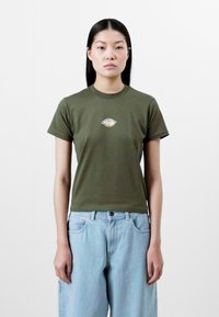 Olive green cotton t-shirt with short sleeves and a crew neckline, featuring a small circular graphic on the chest. Paired with light blue denim jeans.