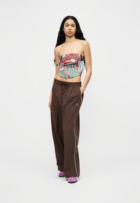 Νεαρή γυναίκα με μακριά σκούρα μαλλιά, που φοράει πολύχρωμο τοπ χωρίς τιράντες, χαλαρό καφέ παντελόνι Puma και μωβ παπούτσια Puma, όρθια με τα χέρια στις τσέπες.