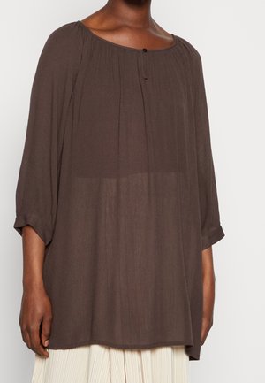 Blouse ample marron foncé à manches trois-quarts avec un bouton unique au col froncé, portée sur un pantalon léger plissé.