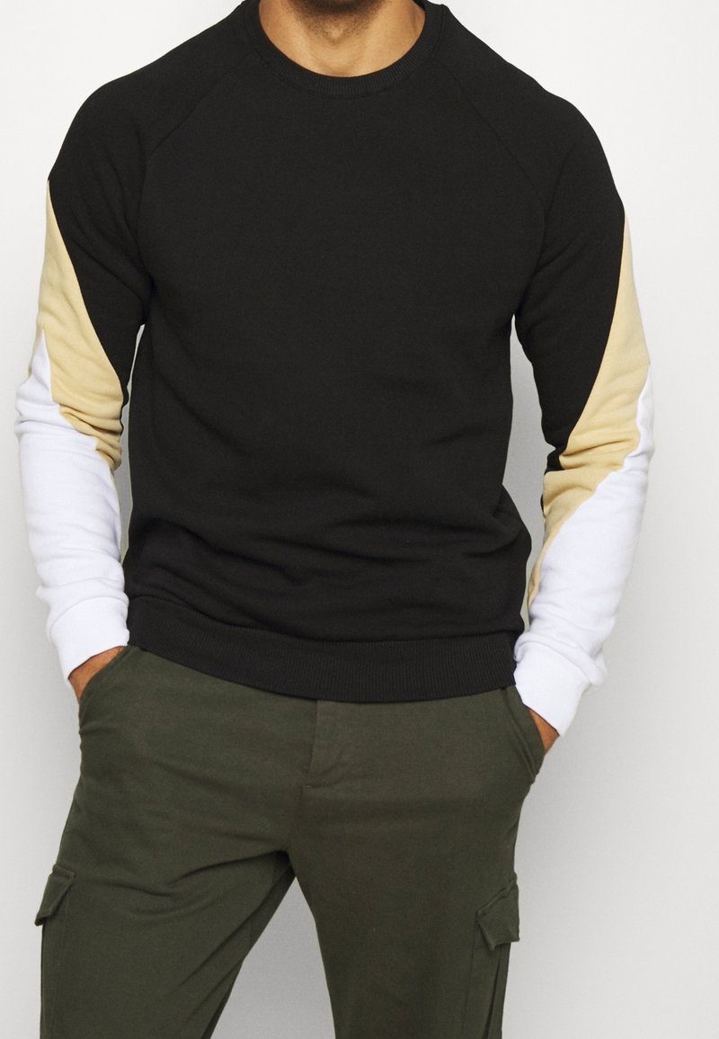 Homme portant un sweat-shirt noir avec des manches beige et blanches en blocs de couleur et un pantalon cargo vert, les mains dans les poches.