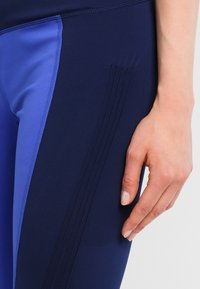 Granatowe legginsy sportowe mają kontrastujące jasnoniebieskie boki oraz dekoracyjne przeszycia na udzie, wykonane z elastycznego materiału.