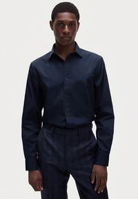 Navyblauw hemd met lange mouwen, knoopsluiting kraag, getailleerde pasvorm, gladde textuur en knopen aan de voorkant. Draagt patroonbroek.