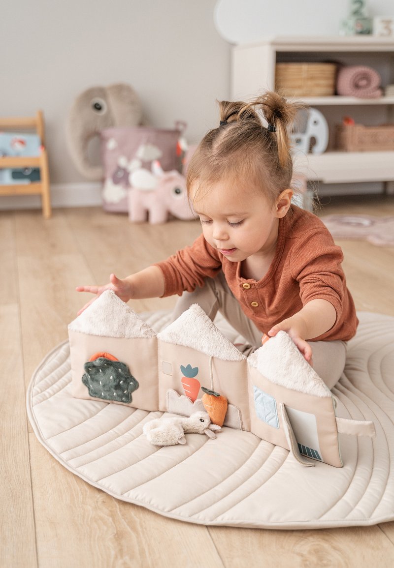 Zacht stoffen speelhuisje in lichtbeige met getextureerde daken, kleurrijke groenteapplicaties en interactieve elementen. Pluche speelgoed vooraan op een ronde mat.