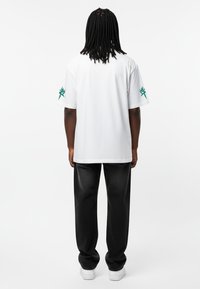 T-shirt blanc à manches courtes avec un détail de logo vert sur la manche gauche, associé à un pantalon noir et des baskets blanches. Texture de tissu lisse.