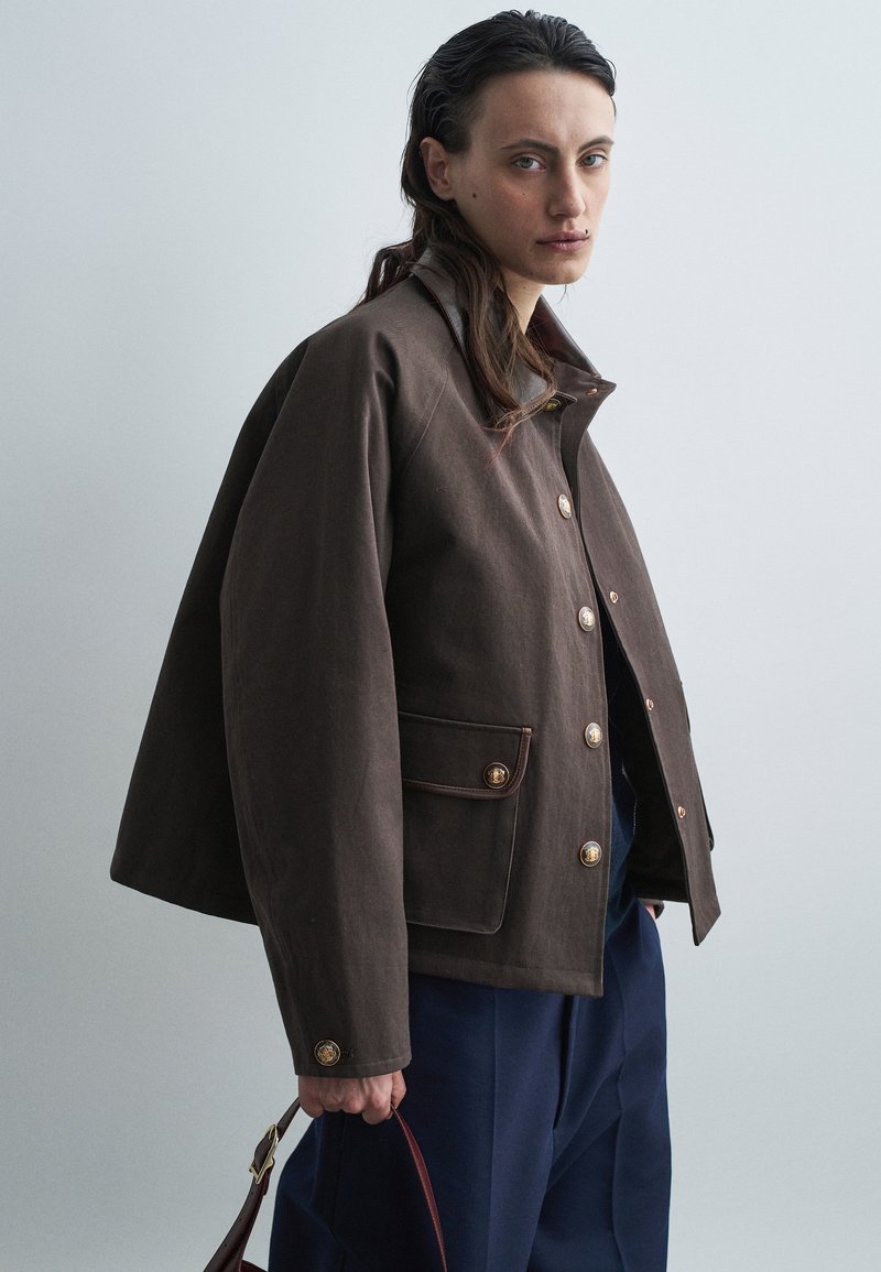 Hnědá zkrácená bunda se zlatými knoflíky, oversized střih, náprsní kapsa s lemováním a klopový límec. Modelka drží burgundskou kabelku.