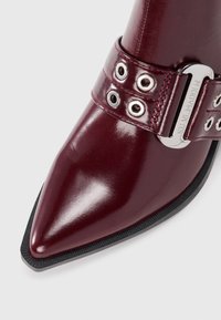 Burgunderrotes Lackleder-Ankle-Boot mit spitzer Zehenpartie, silberner Schnallenhardware und schwarzer strukturiertem Gummisohle mit Ösenakzenten.