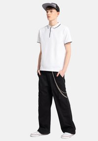 Biała koszulka polo z czarnym wykończeniem, zestawiona z luźnymi czarnymi spodniami cargo z detalami w postaci srebrnego łańcucha. Całość dopełniają białe sneakersy i czapka w kolorze czarnym.