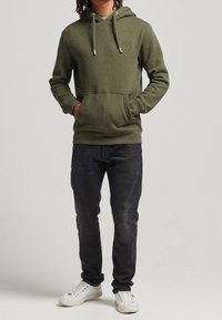 Sudadera verde oliva con capucha ajustable, bolsillo frontal y logo sutil; combinada con jeans negros y zapatillas blancas.