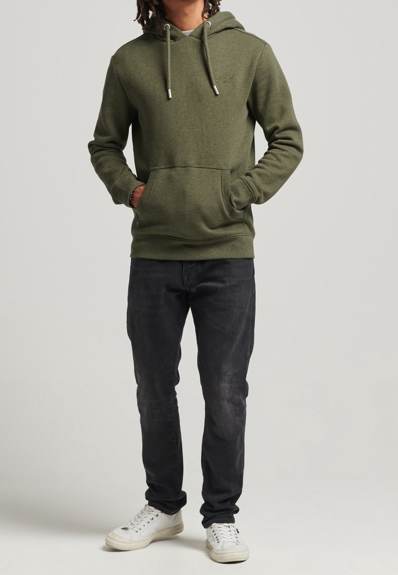 Sudadera verde oliva con capucha ajustable, bolsillo frontal y logo sutil; combinada con jeans negros y zapatillas blancas.