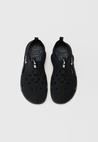 Svarte slip-on sko med teksturert overflate, sirkulære perforeringer og et frontbånd. Nike-logoen er vist i hvitt.