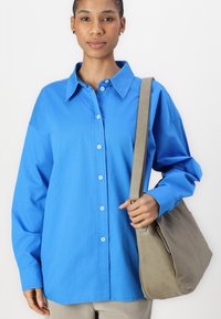 Camicia azzurra chiara con maniche lunghe, dotata di colletto a punte e bottoni bianchi, abbinata a una borsa a spalla grigia.