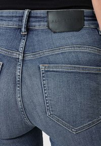 Vaqueros de mezclilla azul con un diseño ceñido, que cuentan con un parche de logo de cuero negro y costuras contrastantes. Los bolsillos traseros tienen una forma clásica cosida.