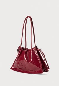 Red patent leather handbag with a triangular shape, gathered top design, and long, slender straps. Features embossed logo on the front.