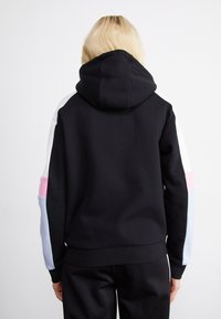 Personne blonde portant un sweat à capuche noir avec des manches color-block en blanc, rose et bleu clair, debout face à un fond blanc.