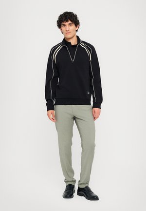Sweatshirt noir avec des lignes blanches, col zippé quart, poignets côtelés, associé à un pantalon vert clair et des chaussures noires.