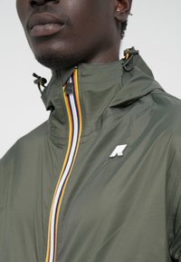 Giacca verde scuro con zip frontale, caratterizzata da una striscia multicolore e un badge logo sul petto. Tessuto leggero e liscio.