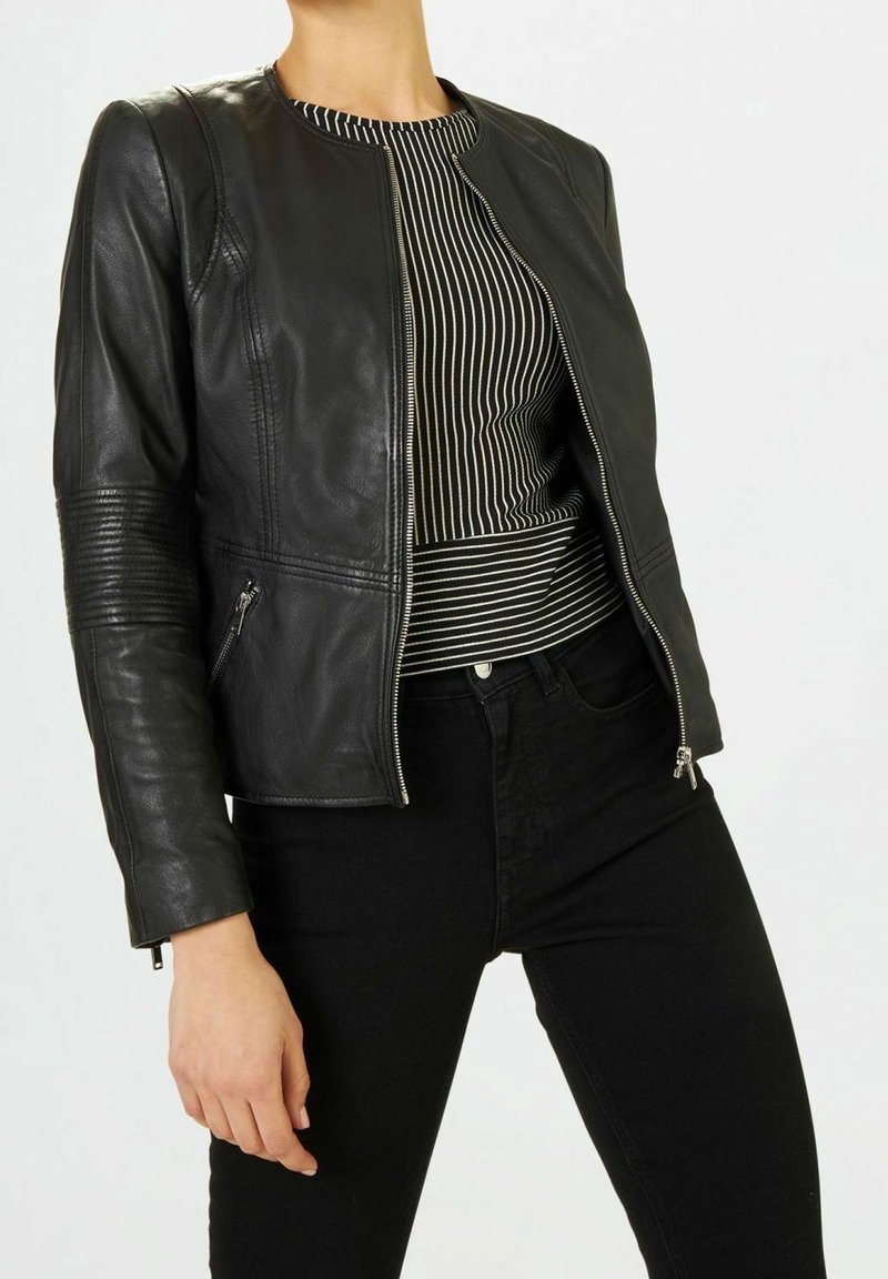Veste en cuir noire à col rond, fermeture éclair devant, détails côtelés aux épaules et deux poches latérales, associée à un haut rayé et un pantalon noir.