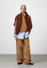 Pull en laine marron porté sur une chemise bleu clair, associé à un pantalon beige large et des chaussures noires ; textures lisses et coupe décontractée.