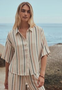 Young woman with long blonde hair wearing beige and striped loose button-up shirt and pants standing by rocky seaside.
