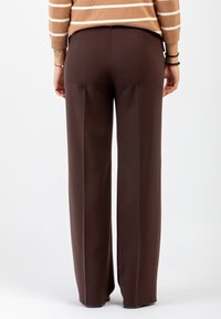 Persona che indossa pantaloni larghi marroni e un maglione beige a righe, in piedi con le mani rilassate ai lati su uno sfondo semplice.