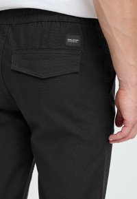 Black pants featuring a textured fabric, elastic waistband, and a back pocket with a tag. The design is minimalist and form-fitting.