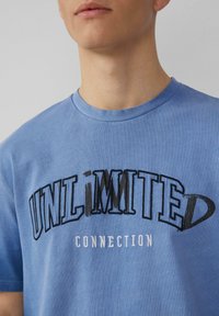 Blauwe katoenen t-shirt met een grafische print van de woorden "ONBEPERKT VERBONDEN" in contrasterend zwart en grijs. Korte mouwen, ronde hals.