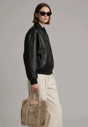 Mujer con chaqueta de cuero negra y gafas de sol, llevando un bolso de pelo sintético beige, de pie frente a un fondo gris liso.