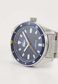 Montre en acier inoxydable ronde avec un cadran bleu marine, des index lumineux, des aiguilles argentées, des accents jaunes et un bracelet en acier inoxydable.