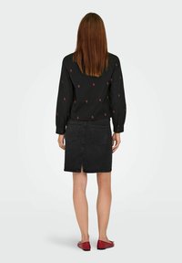 Donna con capelli brown lisci che indossa una blusa nera a maniche lunghe con piccoli motivi floreali rossi, una gonna di denim nera e ballerine rosse, in piedi di spalle.