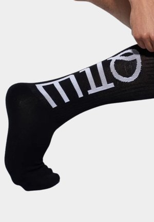 Ręka poprawiająca czarną skarpetę naciągniętą na stopę z dużymi białymi stylizowanymi literami po bokach na jednolitym tle.