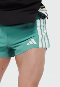 Close-up of a person wearing teal Adidas sports shorts with white three stripes and logo, paired with a black top.