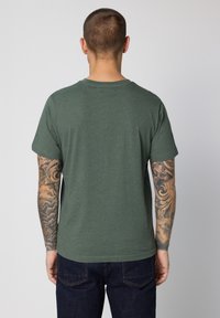 Zelené bavlněné tričko s krátkými rukávy a kulatým výstřihem, hladkou texturou a bez viditelných vzorů, doplněné tmavými džínovými kalhotami.