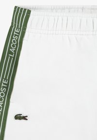 Λευκά βαμβακερά σορτς με πράσινη ελαστική ζώνη μέσης με ριγέ λεπτομέρειες και κεντημένο το λογότυπο της Lacoste στο πλάι.