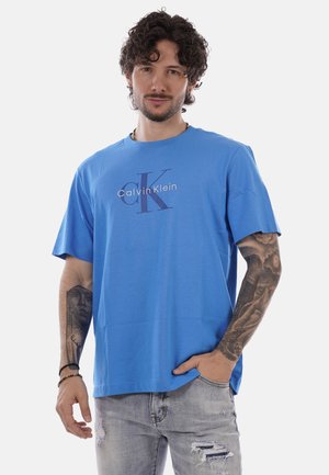 Uomo con capelli ricci e tatuaggi che indossa una maglietta blu Calvin Klein e jeans grigi strappati, posando con una mano in tasca.