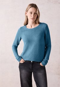 Türkisfarbener, strukturierten Langarmpullover mit Rippenmuster, Rundhalsausschnitt und engen Bündchen, kombiniert mit schwarzen Jeans.