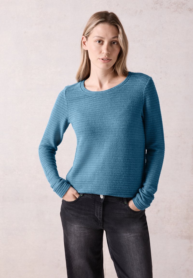 Türkisfarbener, strukturierten Langarmpullover mit Rippenmuster, Rundhalsausschnitt und engen Bündchen, kombiniert mit schwarzen Jeans.