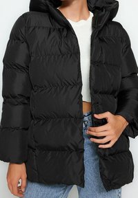 Czarna pikowana kurtka puchowa z kapturem, z haftowanym wzorem, zatrzaskami i szerokimi rękawami. Noszona na białej bluzce i niebieskich dżinsach.