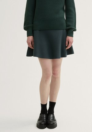 Dunkelgrüner Strickpullover mit gerippten Bündchen, kombiniert mit einem ausgestellten dunkelgrünen Rock und schwarzen Loafers, getragen mit schwarzen Socken.