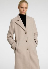 Manteau long beige en mélange de laine douce, doté d'un col à cran, d'une fermeture à cinq boutons et de deux poches latérales. Texture lisse, détails minimaux.