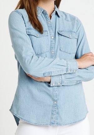 Light blue denim shirt with two chest pockets, button closure, long sleeves, curved hem, and contrasting stitching details.
