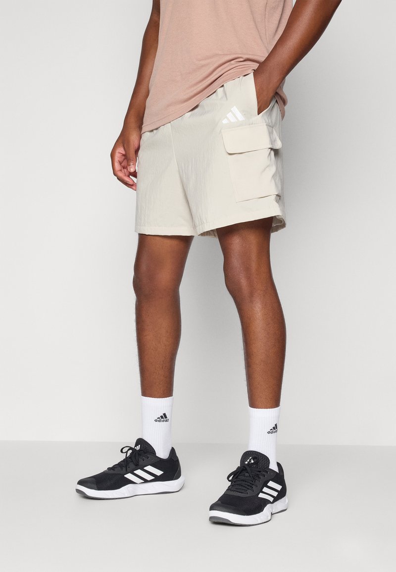 Beige cargo shorts med sidfickor, lättviktigt tyg, i kombination med svarta träningsskor och vita strumpor med tre ränder.