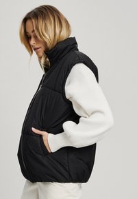 Juoda pūkinė liemenė su aukštu apykakle, užtrauktuku priekyje ir šoninėmis kišenėmis, vilkima ant baltos raukšlėtos megztinio. Dėka lygios apdailos ir paminkštintų dalių.