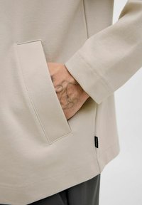 Beige langekout shirt met een gestructureerde stof, met een zijzak en een geëtiketteerde zoom, weergegeven met een hand die gedeeltelijk in de zak is gestoken.